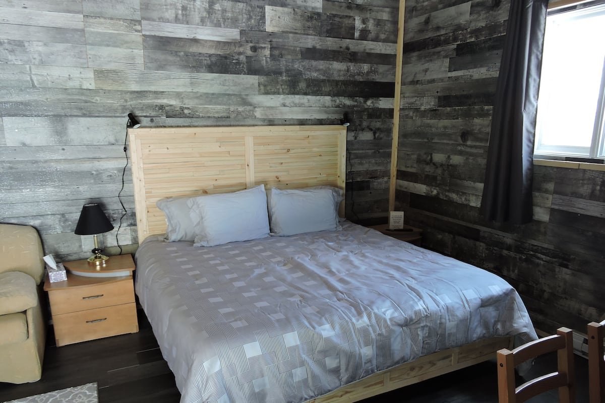 Un dormitorio con cabecera de madera, cama con colcha estampada, mesita de noche con lámpara, una silla y ventana con cortinas.
