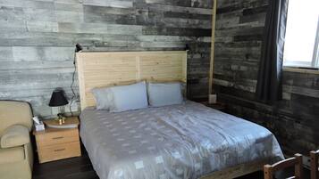 Une chambre à coucher avec une tête de lit en bois, un lit recouvert d’une couvre-lit à motifs, une table de chevet avec une lampe, une chaise et une fenêtre avec des rideaux.