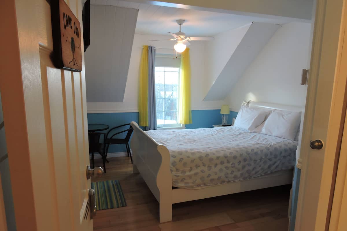 Un dormitorio con cama, escritorio y silla. Se ve una ventana con cortinas y un ventilador de techo.
