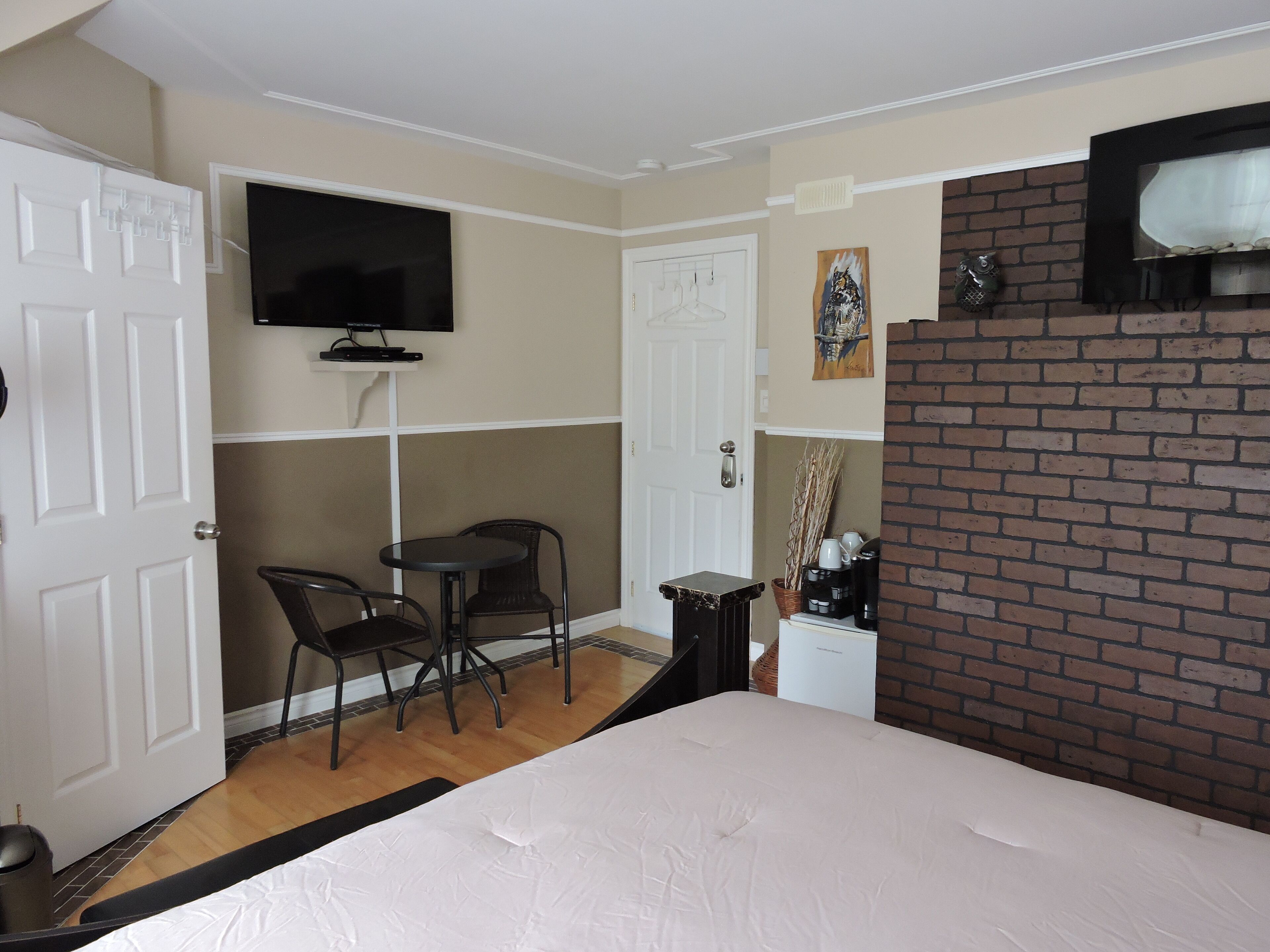 A hotel room with a bed, a TV mounted on the wall, a small table with chairs, a coffee machine, and a brick wall.