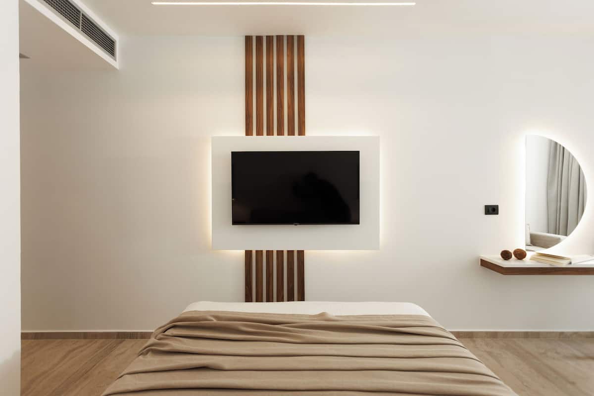 Une chambre à coucher moderne avec un lit, un téléviseur fixé au mur, une table de chevet avec un miroir et un luminaire fixé au mur.