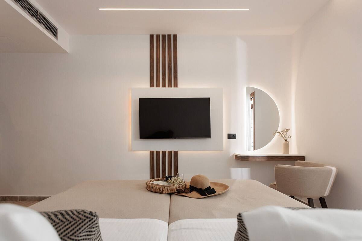Une chambre d’hôtel moderne dotée d’un grand téléviseur à écran plat fixé au mur, d’un miroir rond au-dessus d’une coiffeuse et d’un coin salon confortable avec un canapé et des fauteuils.