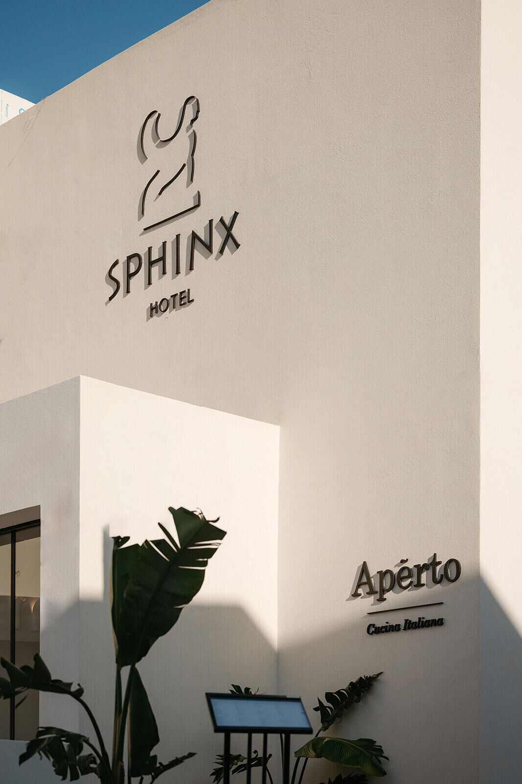 The exterior of the Sphinx Hotel with a large plant in the foreground and a sign for the Italian cuisine restaurant, Aperto.