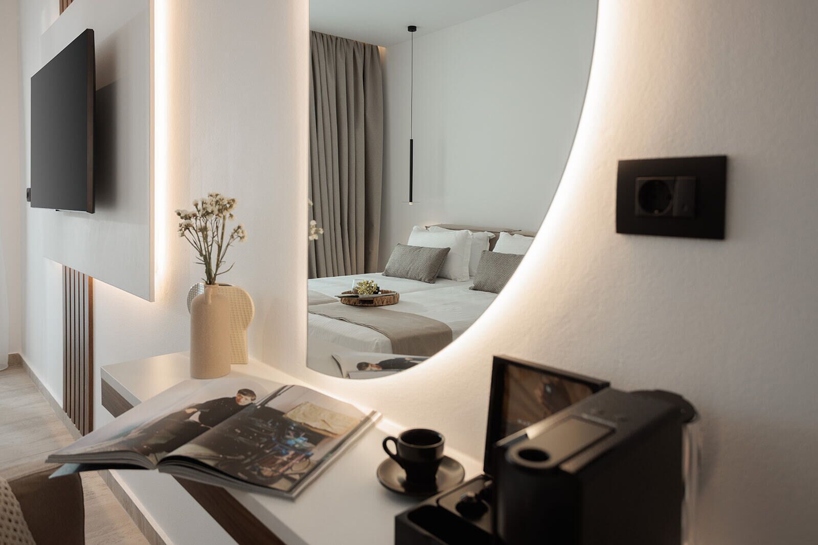 A modern hotel room with a large mirror reflecting the bed and curtains, a flat-screen TV mounted on the wall, a bedside table with a vase of flowers, and a magazine.