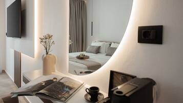 A modern hotel room with a large mirror reflecting the bed and curtains, a flat-screen TV mounted on the wall, a bedside table with a vase of flowers, and a magazine.