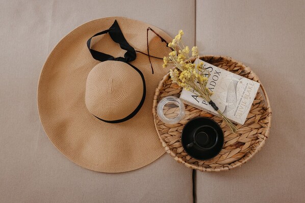 Un sombrero de paja con un lazo negro, una taza negra, un frasco de vidrio, flores secas y un libro con el título 'Anything by Olive Blake' sobre una bandeja tejida.