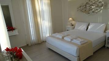 Quarto de hotel com uma cama grande, uma mesinha de cabeceira com abajur, um espelho e uma janela com cortinas leves.