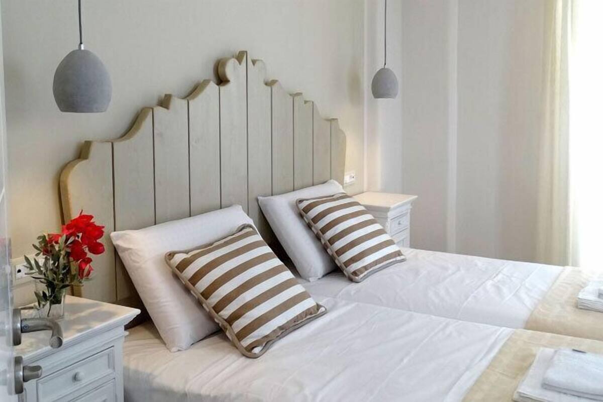 Une chambre d’hôtel avec deux lits, du linge de lit blanc, des oreillers à rayures, une table de chevet avec une lampe et un vase rempli de fleurs rouges.