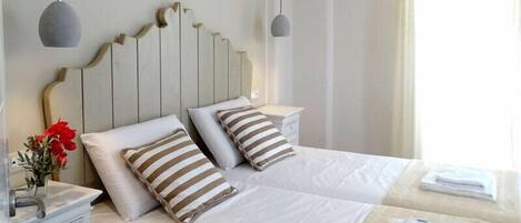Hotellihuone, jossa on kaksi sänkyä, valkoiset vuodevaatteet, raidalliset tyynyt, yöpöytä lampun kanssa ja maljakko, jossa on punaisia kukkia.