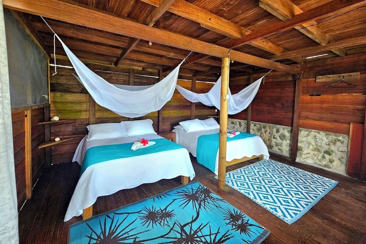 Eine Holzhütte mit zwei Betten, jedes mit einem weißen Moskitonetz und türkisblauen Bettdecken.