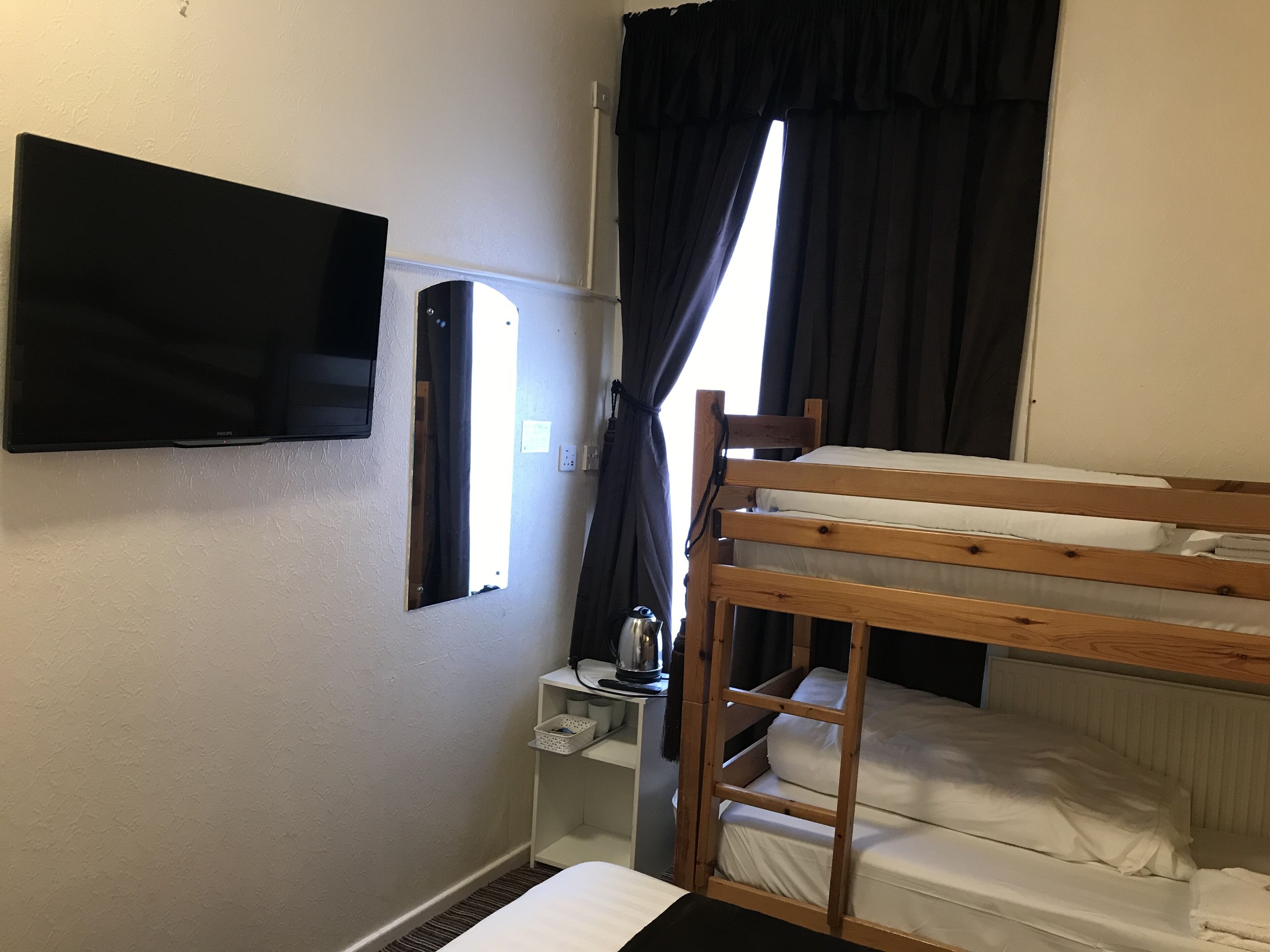 A bunk bed room with a television mounted on the wall, a small table with a kettle, and a window with curtains.