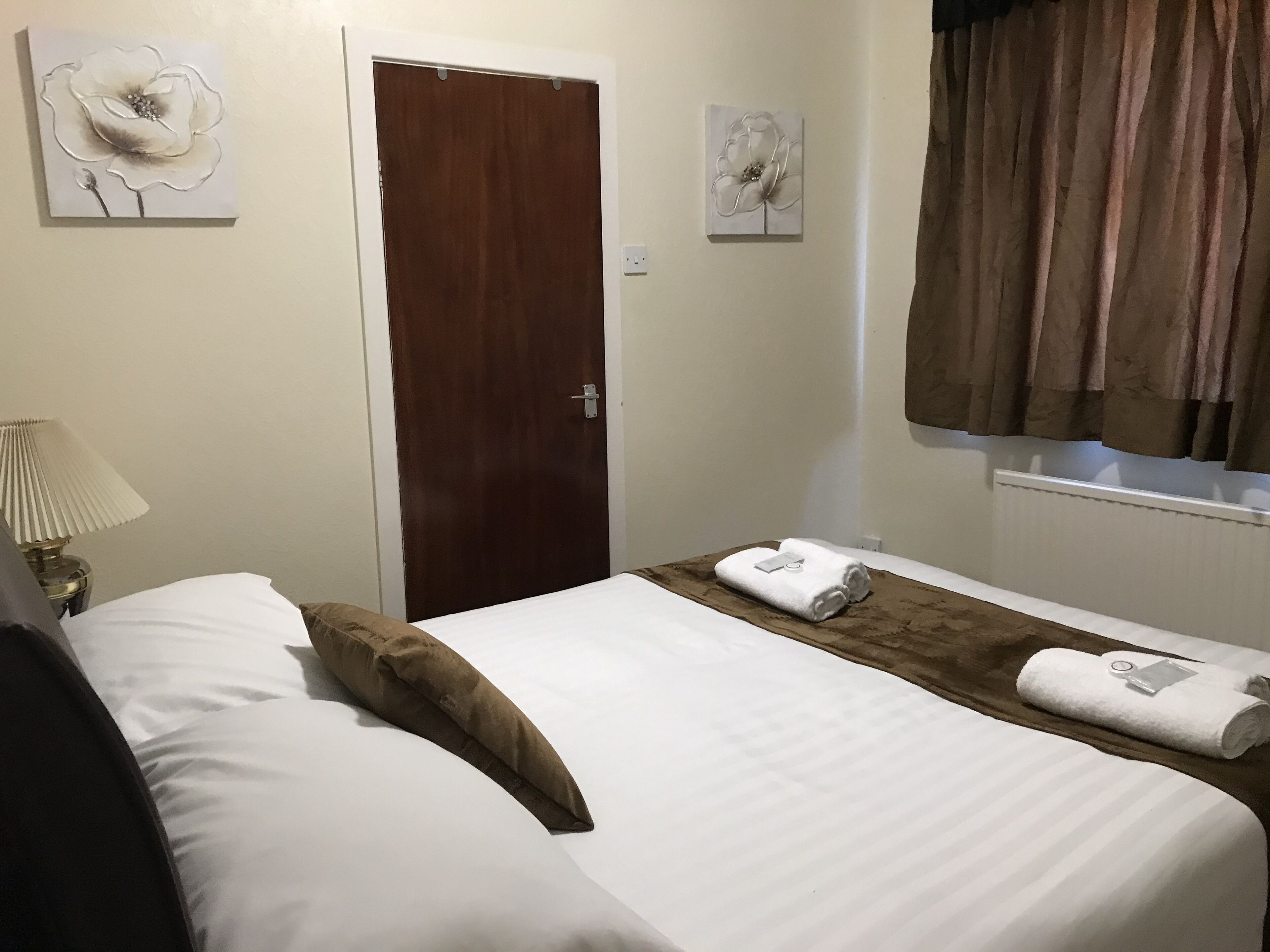 A hotel room with a bed, a nightstand with a lamp, a wooden door, and two wall-mounted flower paintings.
