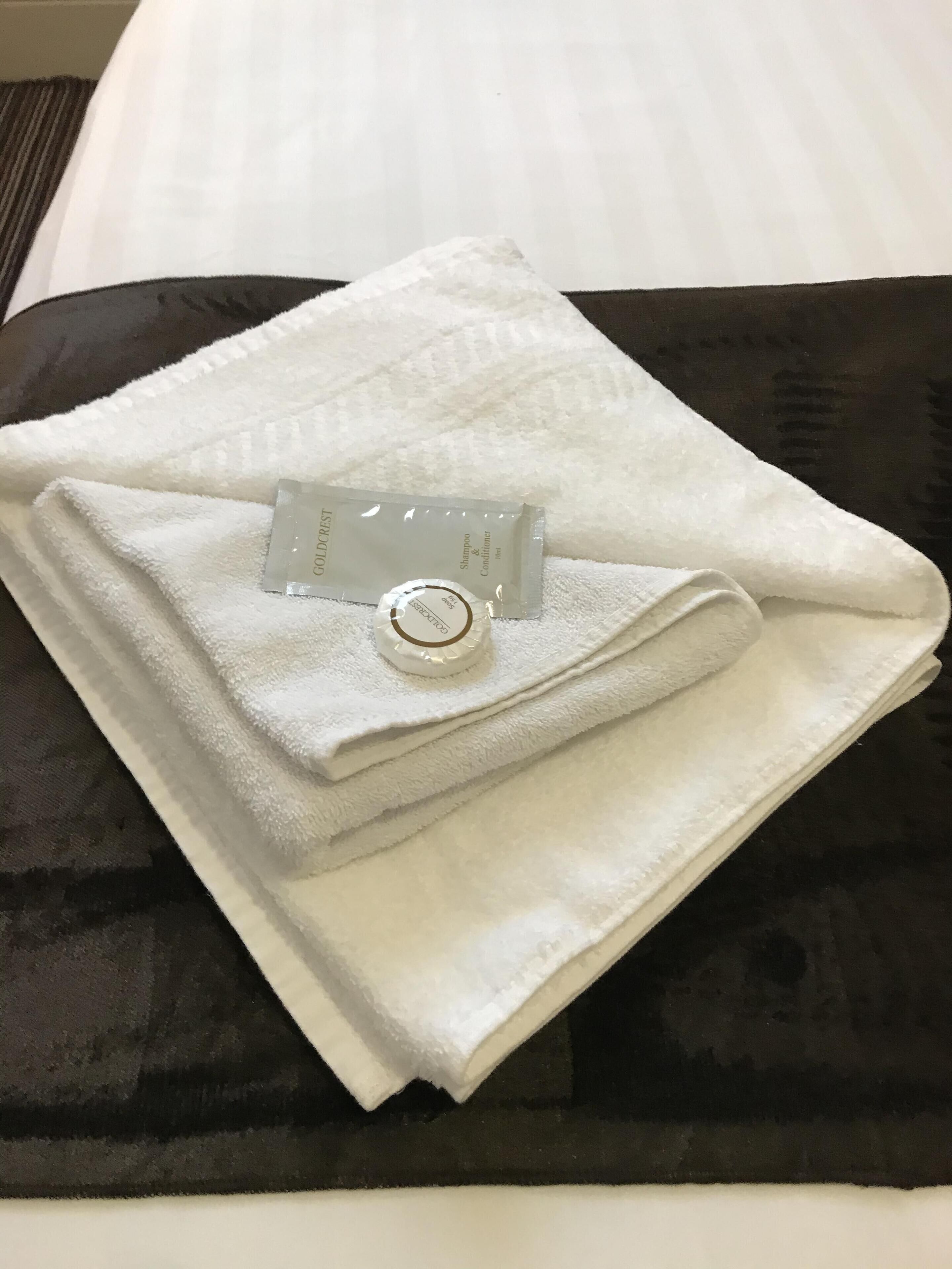 Une serviette blanche soigneusement pliée, accompagnée d’une bouteille de shampoing et d’après-shampoing Goldcrest à l’étiquette dorée, est posée sur un coussin noir.