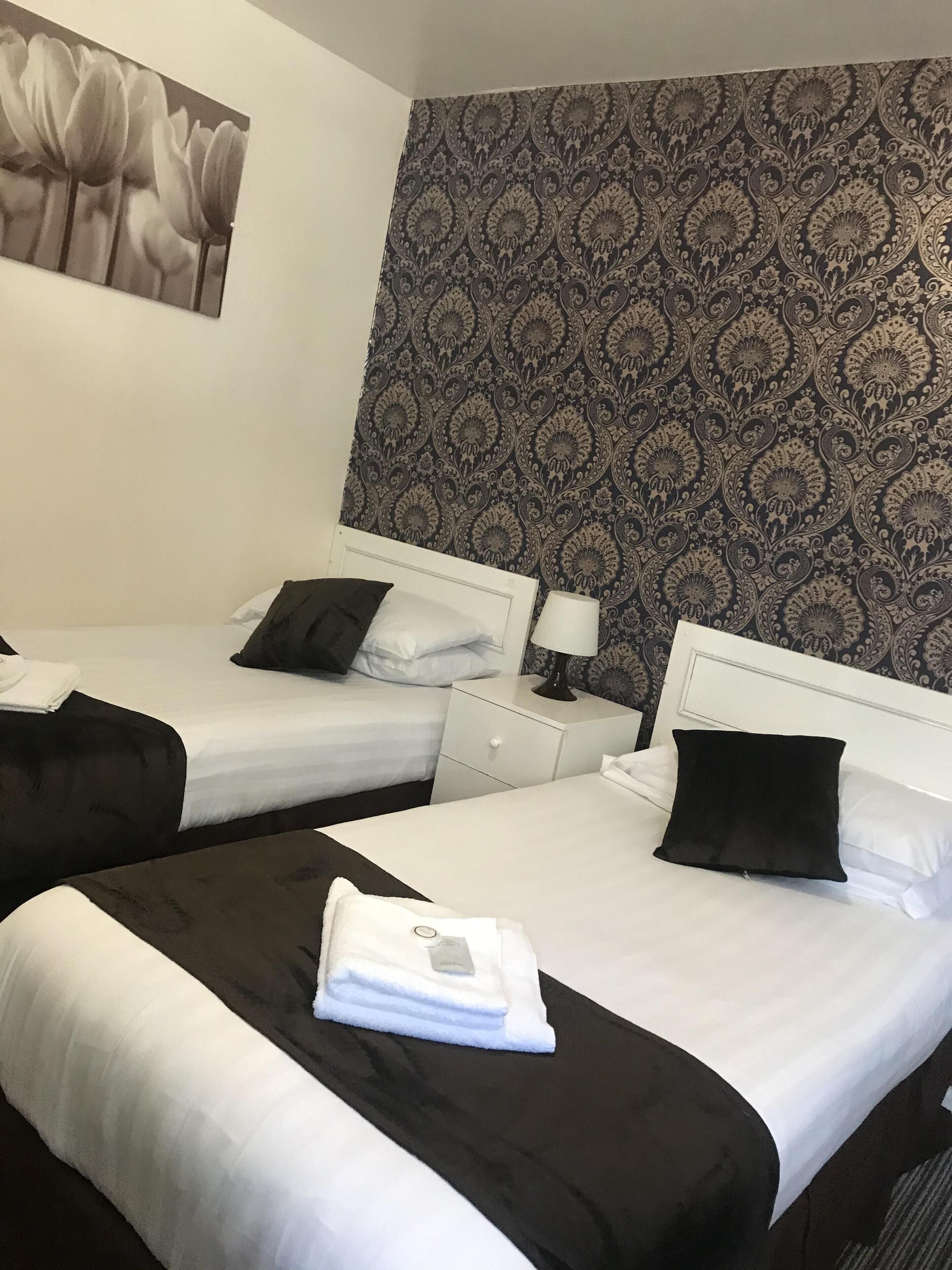 Habitación de hotel con dos camas, ropa de cama blanca, almohadas negras, una mesita de noche con lámpara y un papel tapiz estampado.
