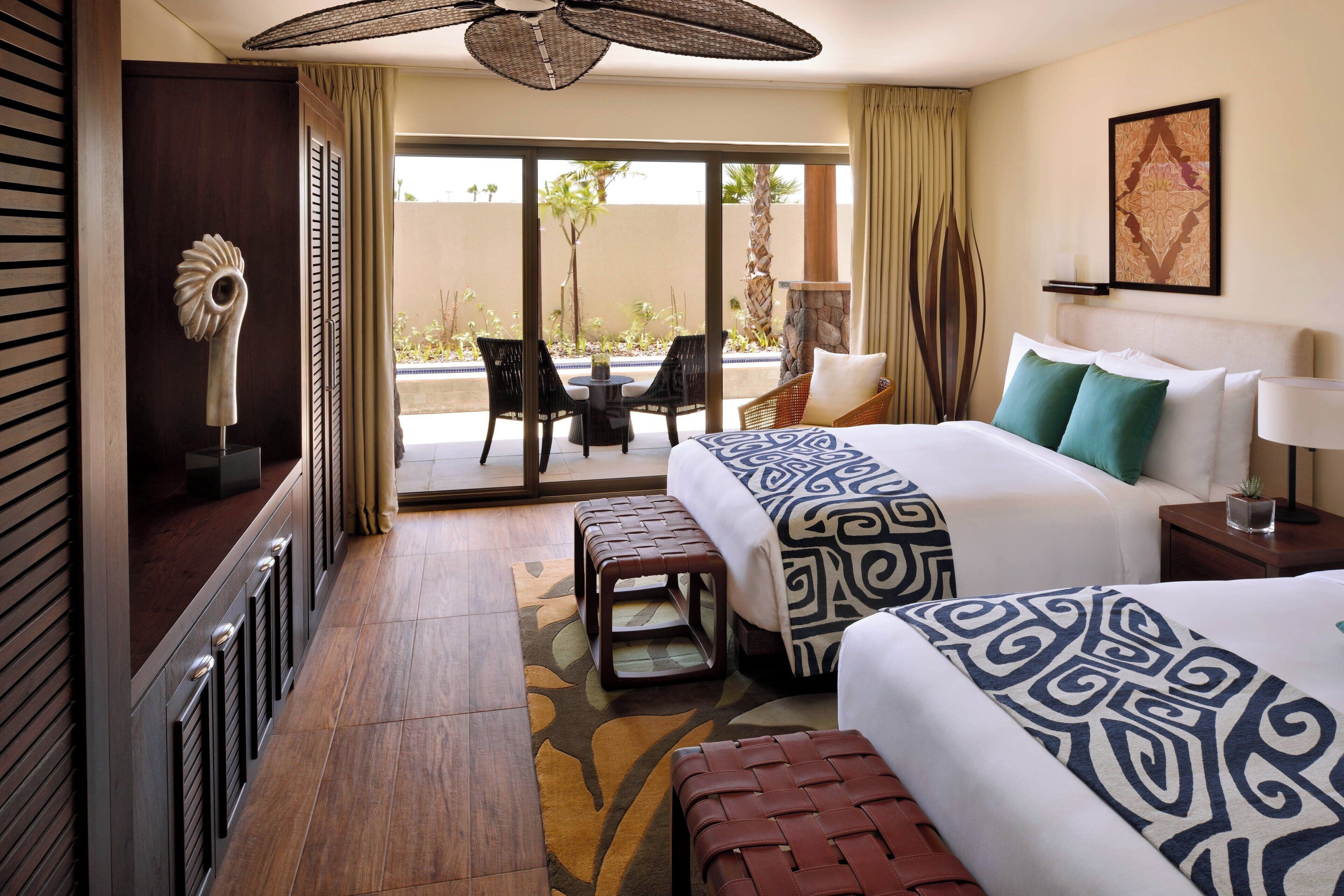 A hotel room with a large bed, a small table, two chairs, a bench, and a view of a pool and palm trees.