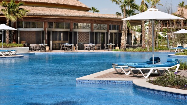 A pool area with lounge chairs, umbrellas, and a building with a thatched roof. - Lapita, Dubai Parks and Resorts, Autograph Collection (Dubai)