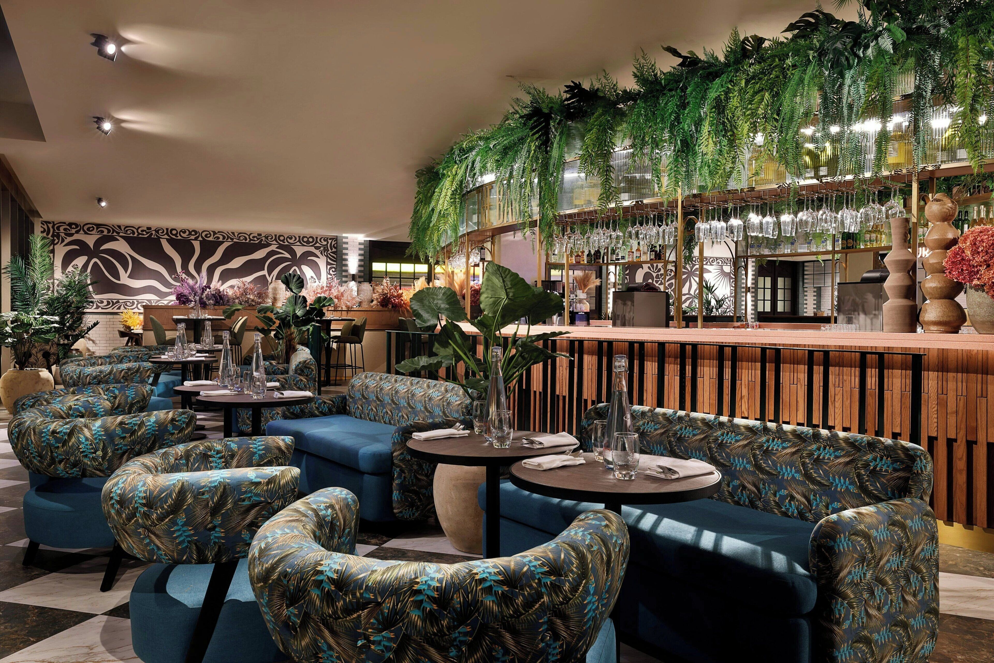 A lounge area with blue upholstered sofas, round tables, and a bar area with hanging plants and glassware.