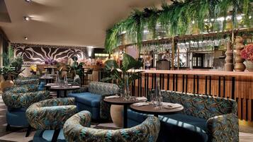 A lounge area with blue upholstered sofas, round tables, and a bar area with hanging plants and glassware.