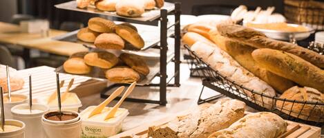 Variedad de panes y pasteles expuestos, incluyendo baguettes y bollos, junto con recipientes de untables y mantequilla.