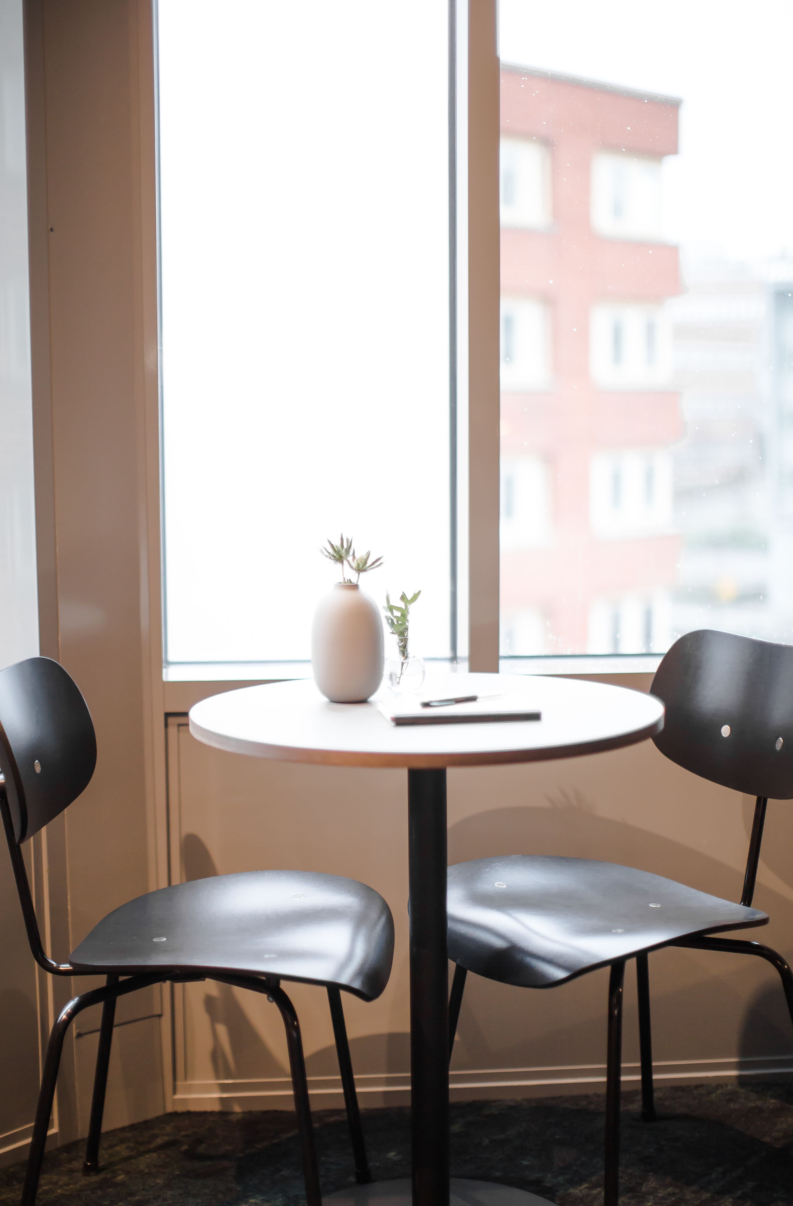 A room with a round table, two chairs, a vase with a plant, and a view of buildings through a window.