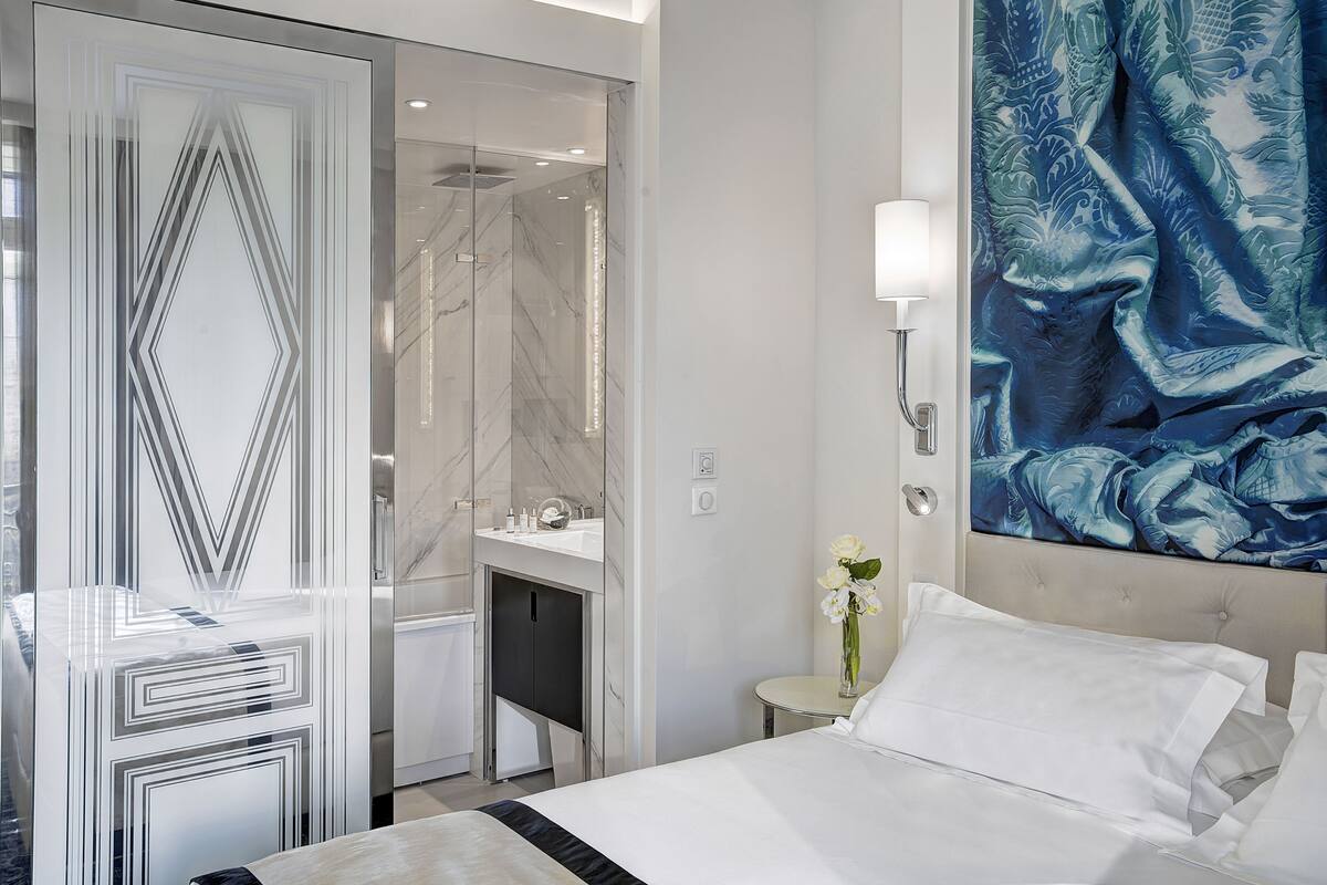 A modern hotel room with a large bed, a glass partition wall, a bathroom with a marble shower, and a large floral artwork on the wall.