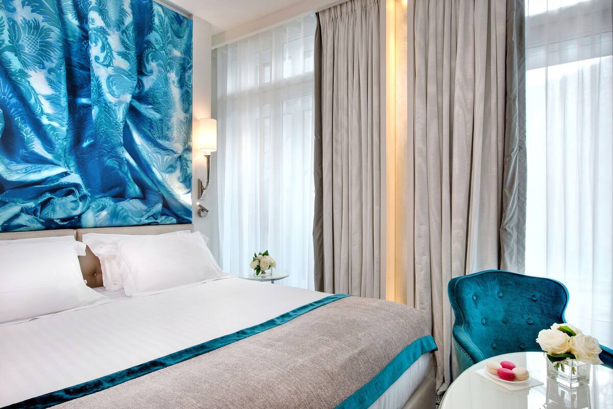 A hotel room with a bed, a blue upholstered chair, a small table with a vase of flowers, and a view of the outside through sheer curtains.