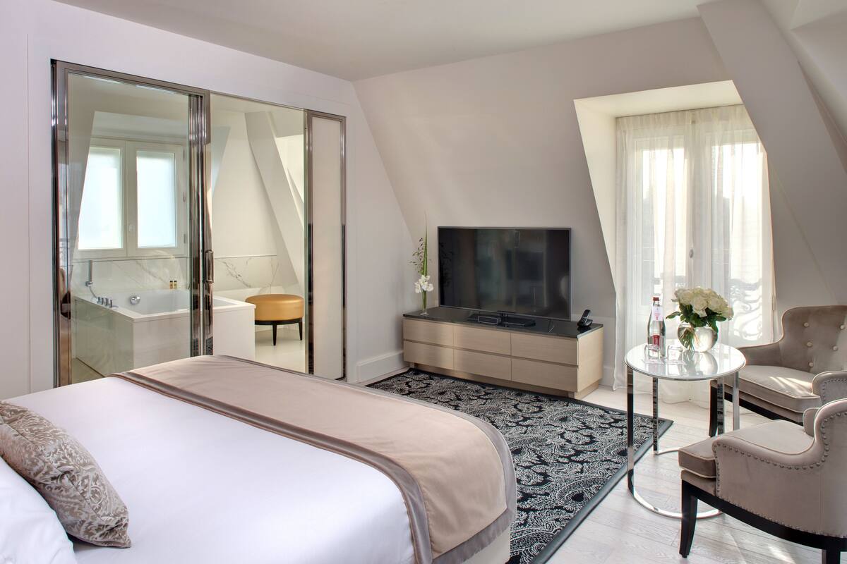 A modern hotel room with a large bed, a TV, a sofa, and a bathroom visible through a sliding door.