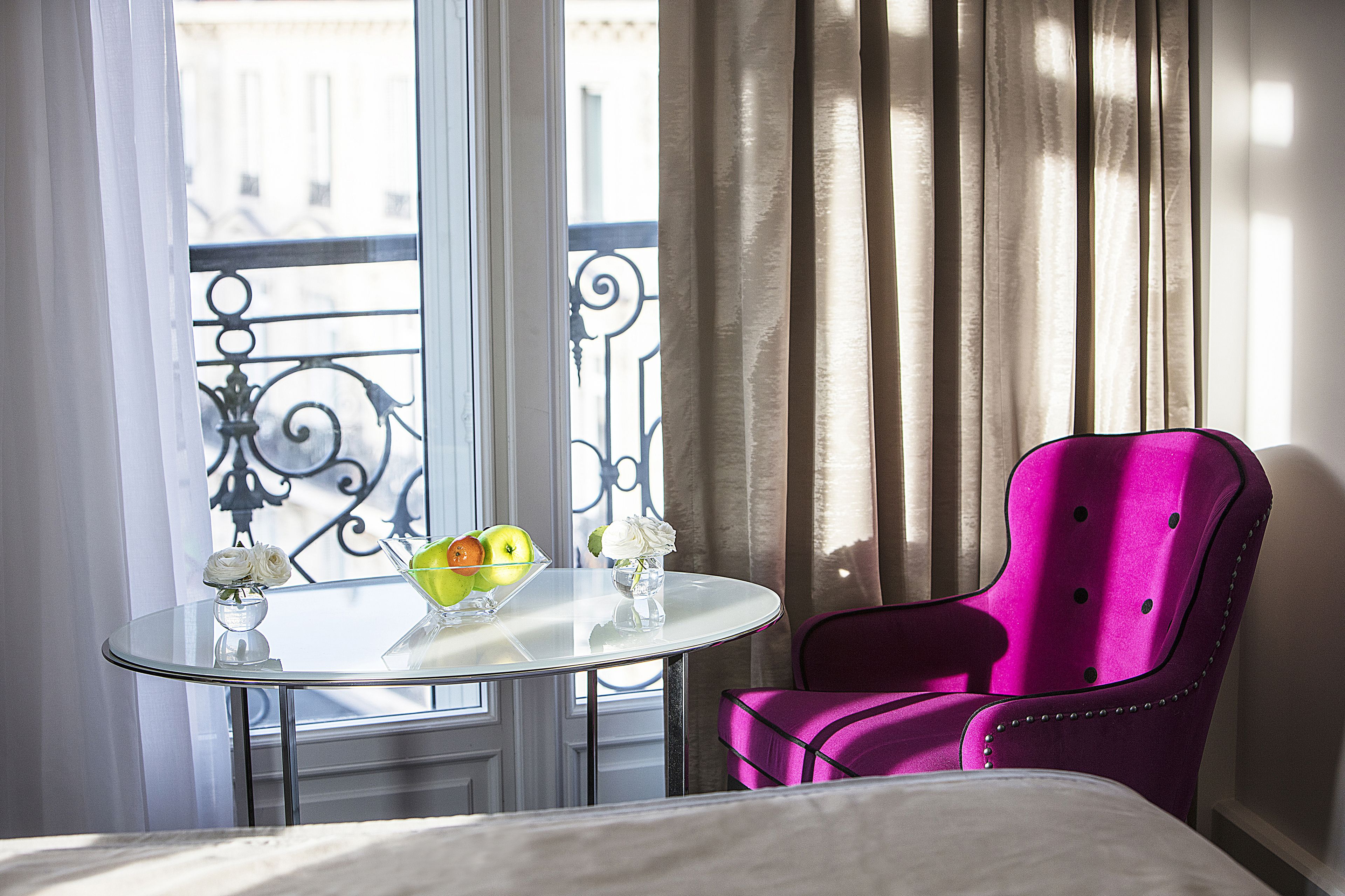 A hotel room with a bed, a round glass table with a bowl of fruit and a vase of flowers, a pink chair, and a view of the outside through a window with curtains.