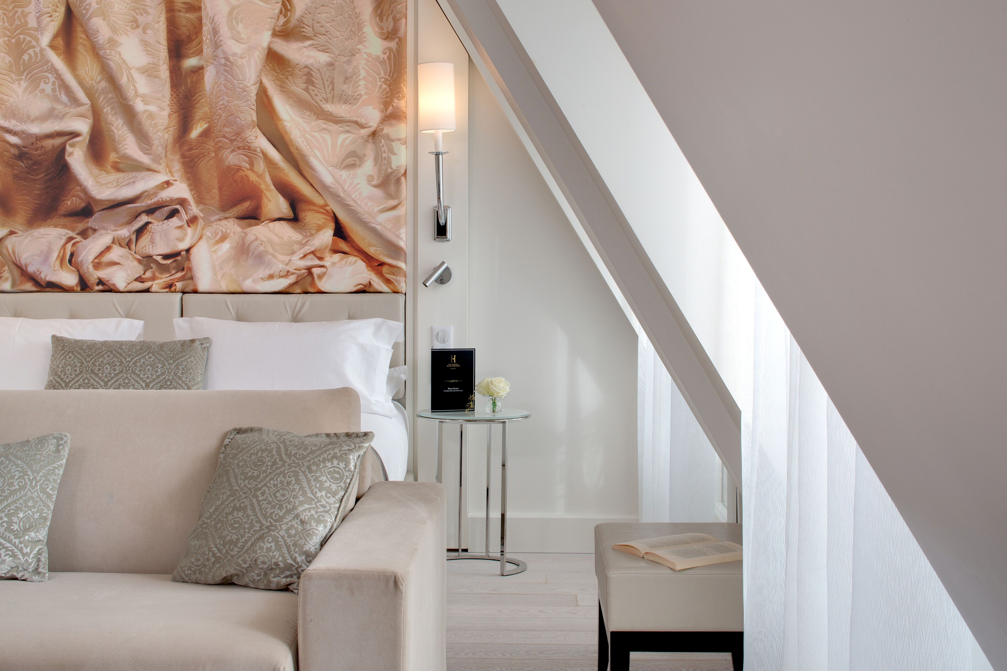 Une chambre à coucher moderne avec un grand lit, une petite table sur laquelle se trouvent un livre et un vase, et une lampe fixée au mur.