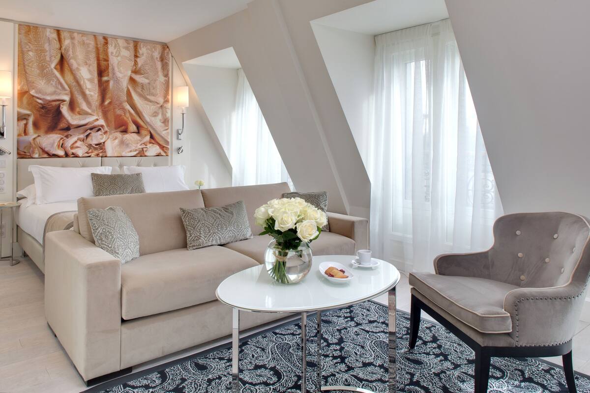 A modern hotel room with a large bed, a sofa, a coffee table, and a chair. There is a painting on the wall and a window with curtains.