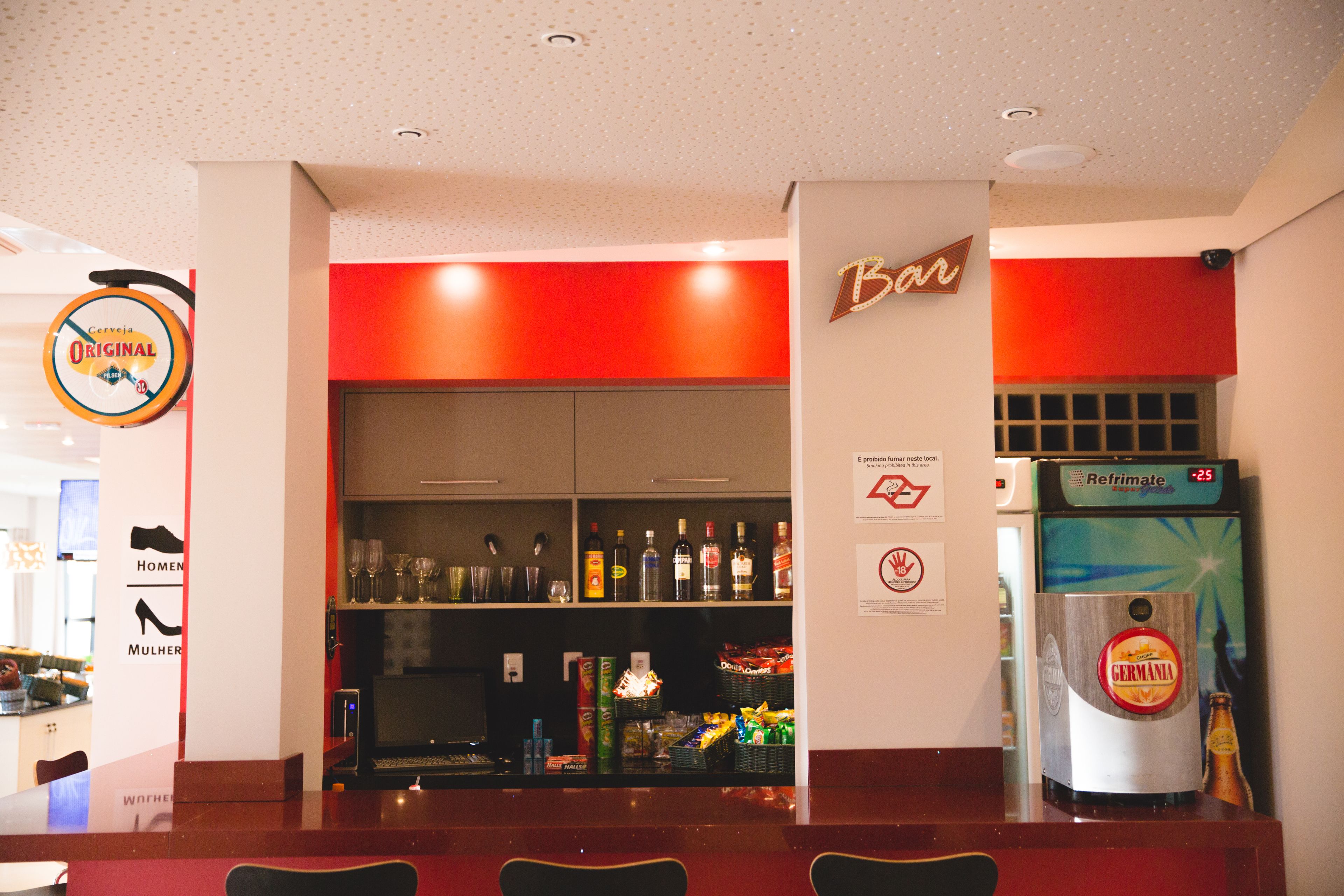 A bar area with a refrigerator stocked with beverages, shelves with bottles, and a sign indicating the bar's name.