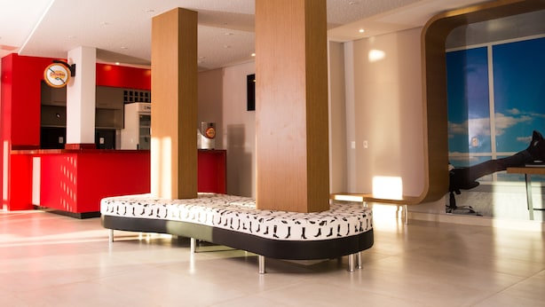 A modern lobby with a red reception desk, a seating area with a patterned bench, and a large window with a view of the sky. - ibis Styles Birigui (Birigui)