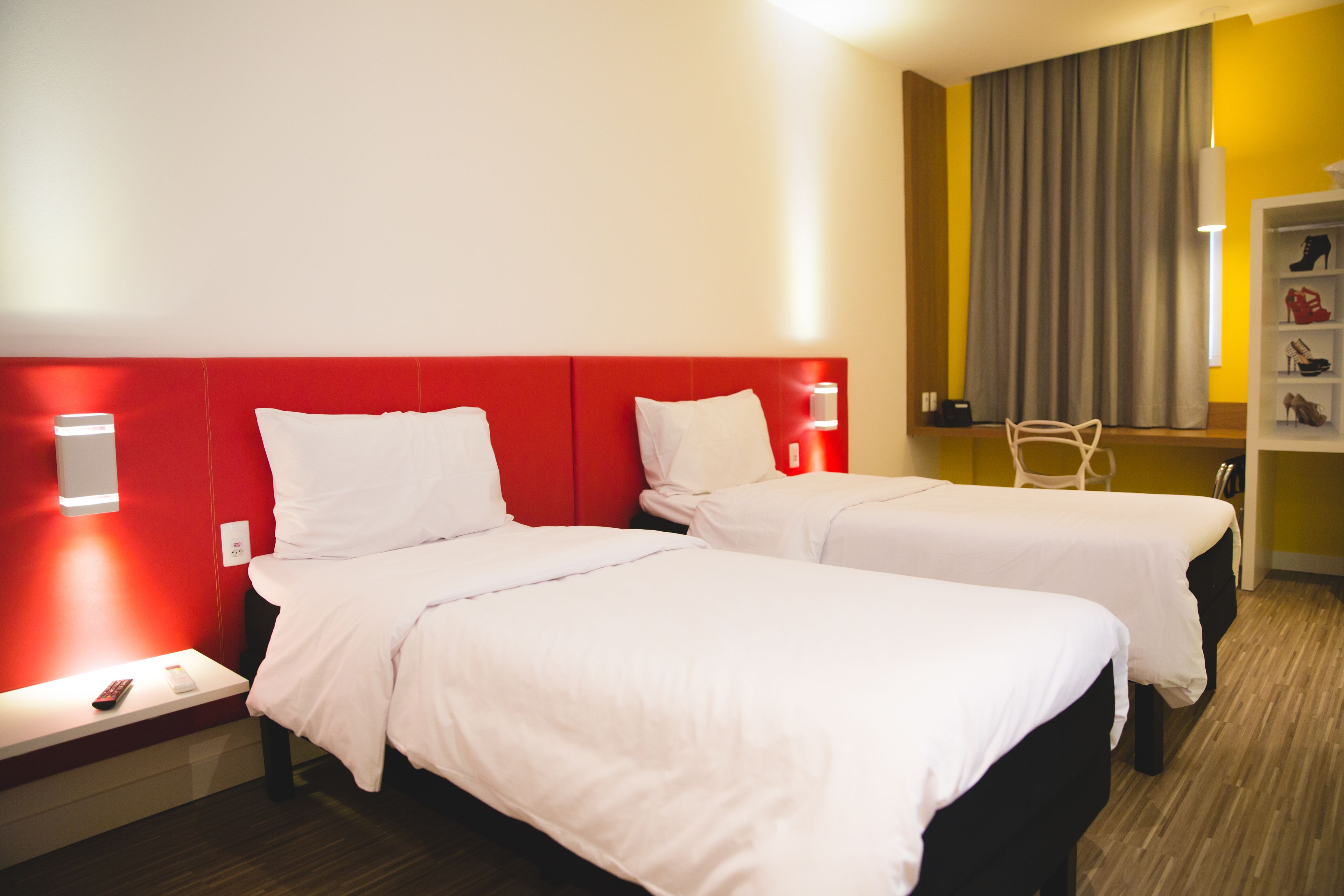 Habitación de hotel con dos camas, cabecera roja, ropa de cama blanca, mesita de noche con teléfono y zapatero.