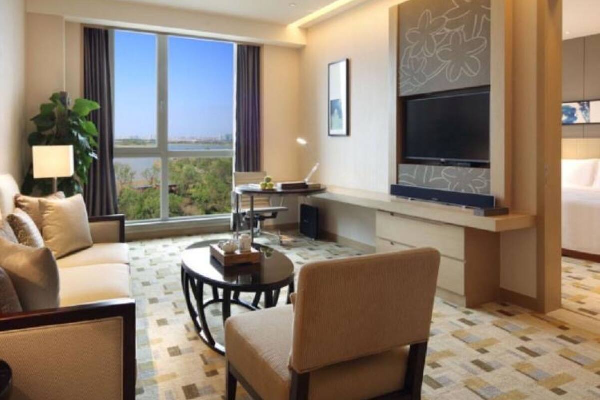 A hotel room with a sofa, armchair, dining table, and a large window offering a city view.