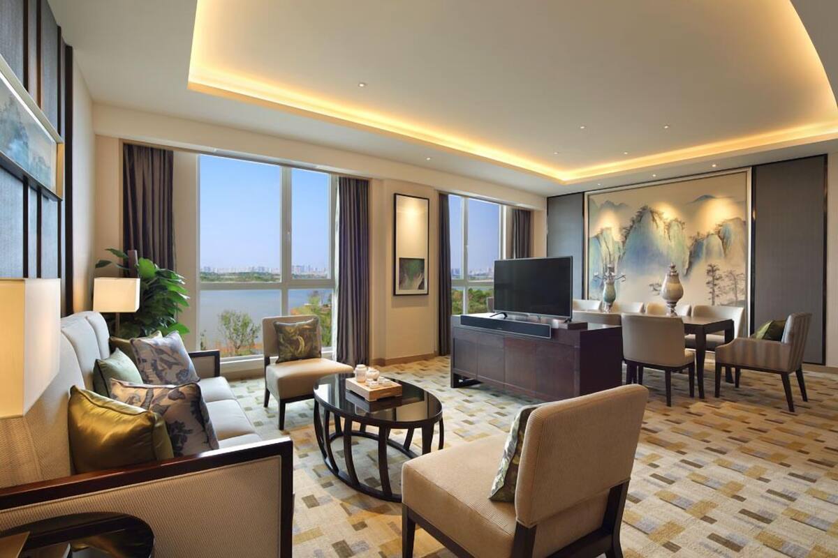 A modern hotel room with a large flat-screen TV, a dining area, and a view of the cityscape through large windows.
