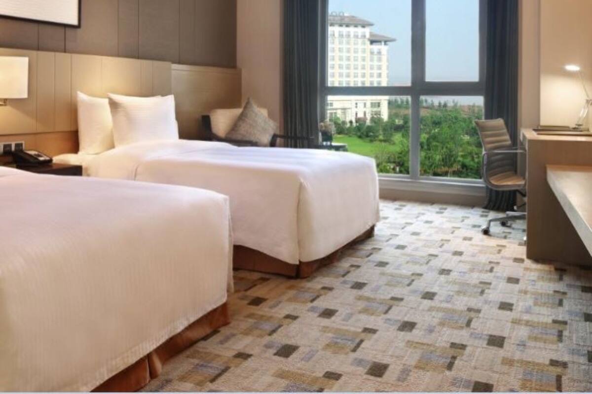 A hotel room with two beds, a desk, and a large window offering a city view.