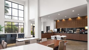 A modern kitchen with a central island, dining area with tables and chairs, and large windows providing natural light.