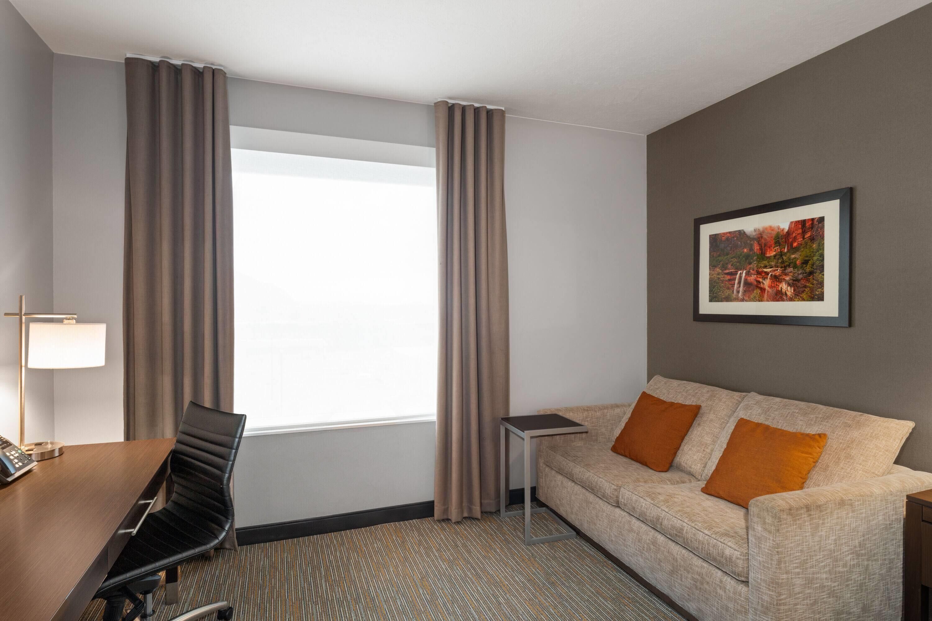 A hotel room with a bed, a desk with a lamp, a chair, a small table, and a sofa with two cushions.