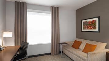 A hotel room with a bed, a desk with a lamp, a chair, a small table, and a sofa with two cushions.