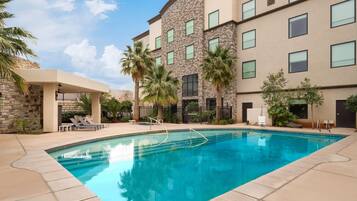 A swimming pool with lounge chairs and palm trees, surrounded by a building with stone and beige walls and multiple windows.