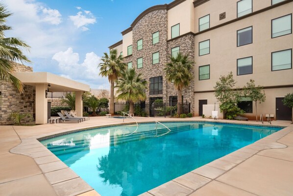 A swimming pool with lounge chairs and palm trees, surrounded by a building with stone and beige walls and multiple windows.