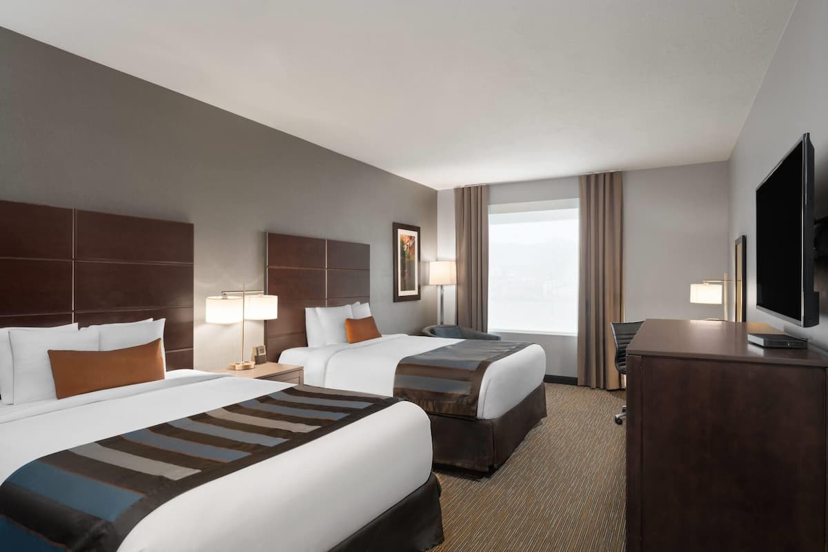 A hotel room with two beds, a desk, a TV, and a large window with curtains.