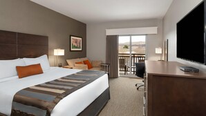 A hotel room with a large bed, a desk with a TV, a balcony with a view, and a painting on the wall.