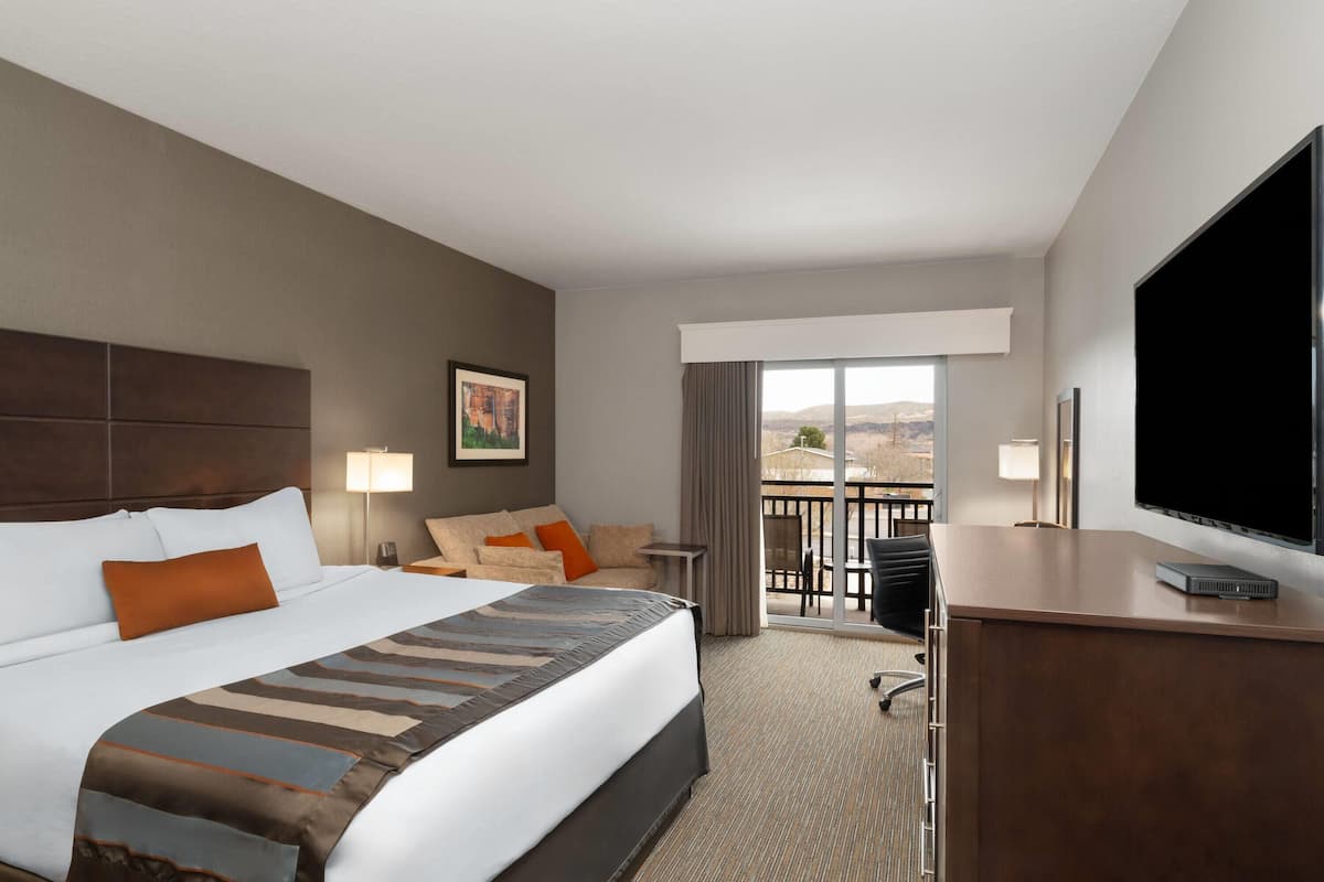 A hotel room with a large bed, a desk with a TV, a balcony with a view, and a painting on the wall.