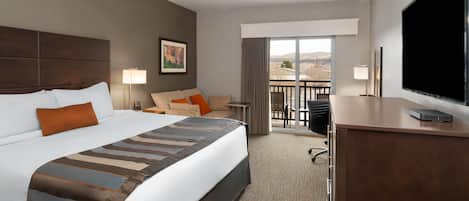Habitación de hotel con una cama grande, un escritorio con televisor, un balcón con vistas y un cuadro en la pared.