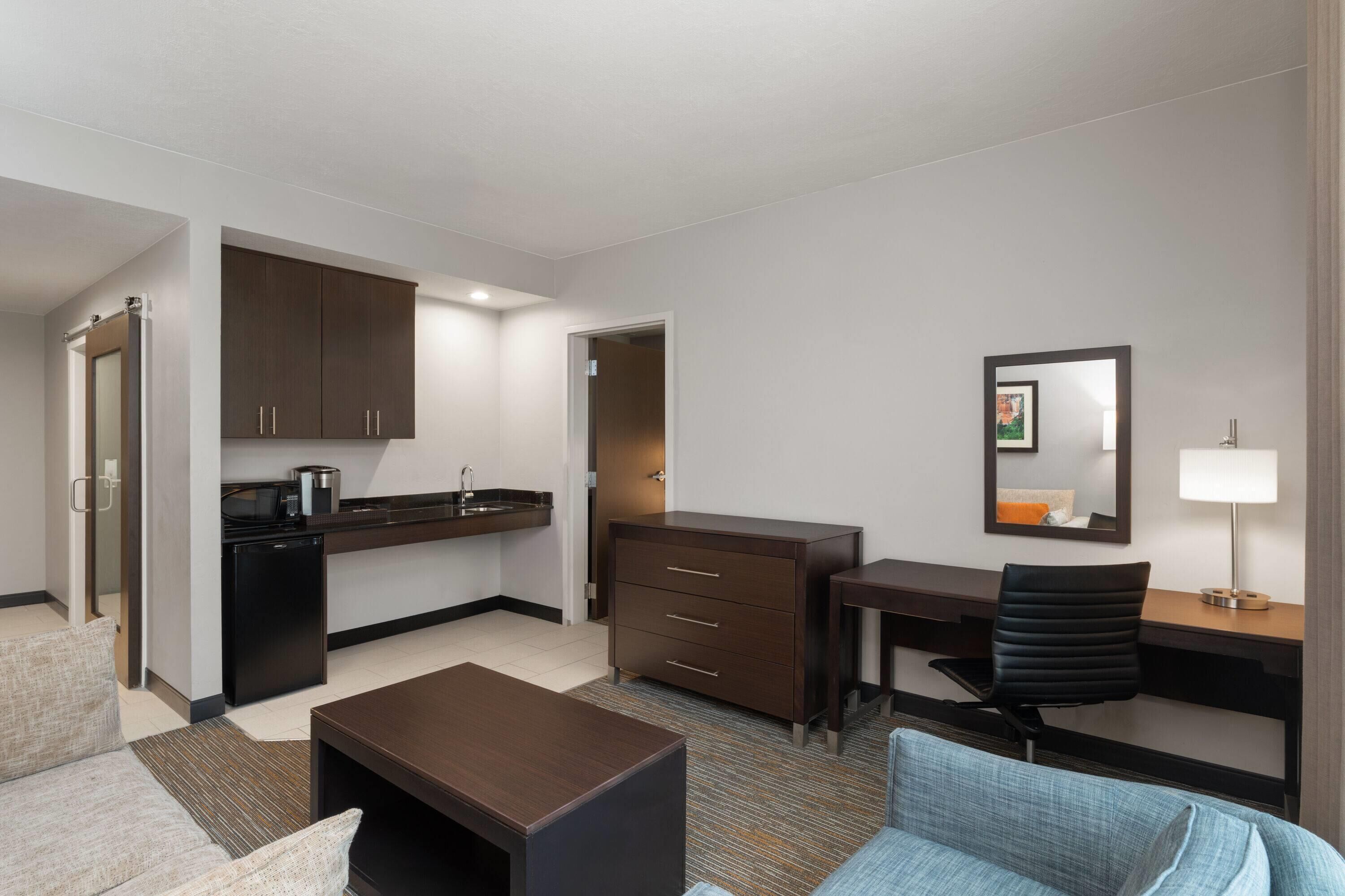 A compact hotel room with a kitchenette, a desk, a chair, a sofa, and a small table.