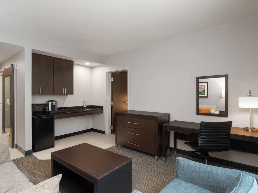 Habitación de hotel compacta con kitchenette, escritorio, silla, sofá y una pequeña mesa.