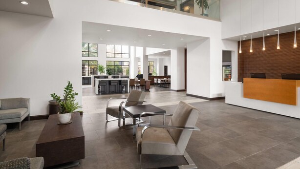 A spacious lobby with a high ceiling, a reception desk, seating area, and a kitchen in the background.