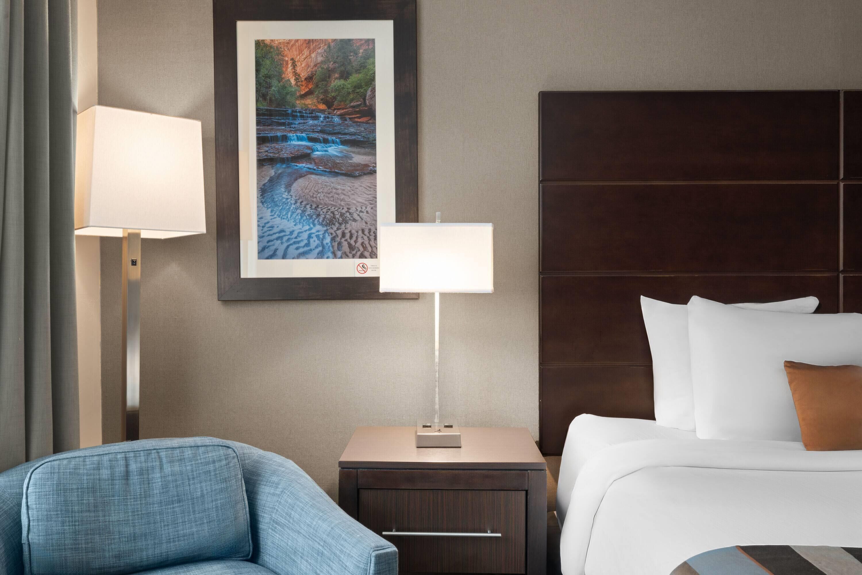 A hotel room with a bed, a nightstand with a lamp, a chair, and a framed picture on the wall.