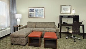 A hotel room with a sectional sofa, two ottomans, a desk with a chair, a lamp, and a framed picture on the wall. - Staybridge Suites Houston - Medical Center by IHG (Houston)